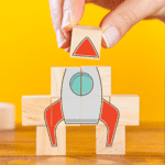 A series of stacked wooden blocks forming the image of a rocket ship in the process of being built by a woman's hand.